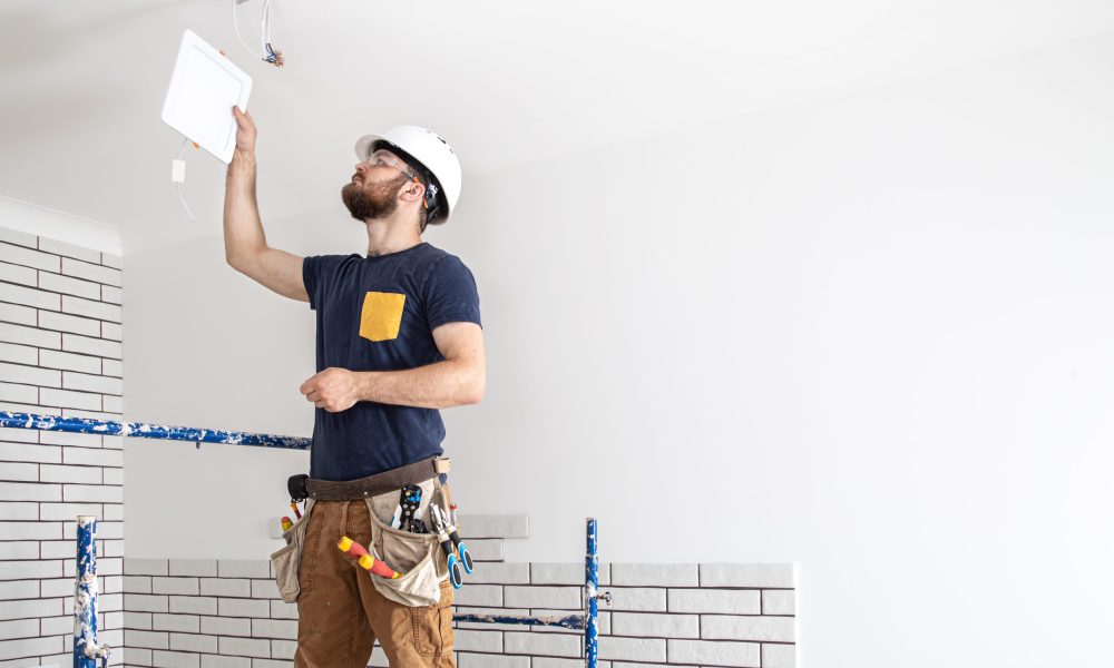 Electrician Builder with beard worker in a white helmet at work, installation of lamps at height. Professional in overalls with a drill on the background of the repair site.